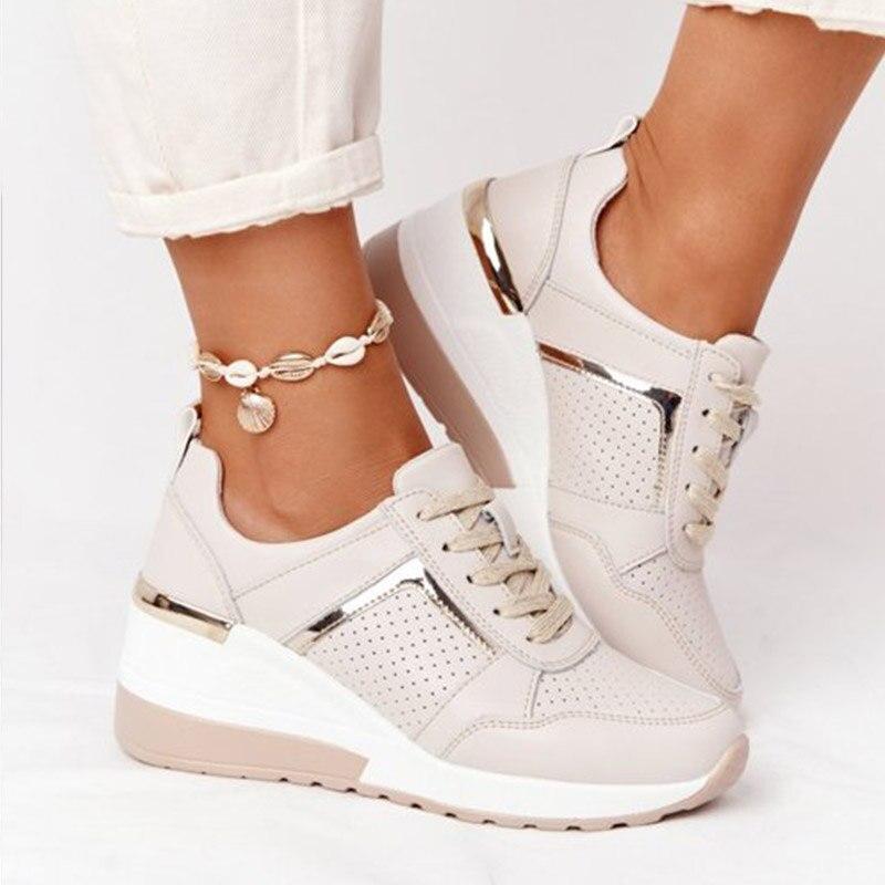 Beige orthopedic sneakers with wedge heel, metallic accents, and perforated design, worn with white pants and shell anklet. Comfortable footwear.