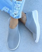 Gray slip-on sneakers with white soles, featuring a breathable mesh design. Paired with ripped light blue jeans on a concrete surface. Fashionable casual footwear.