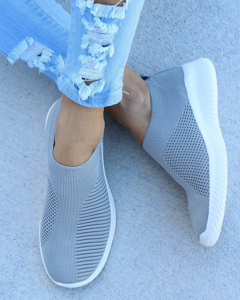 Gray slip-on sneakers with white soles, featuring a breathable mesh design. Paired with ripped light blue jeans on a concrete surface. Fashionable casual footwear.