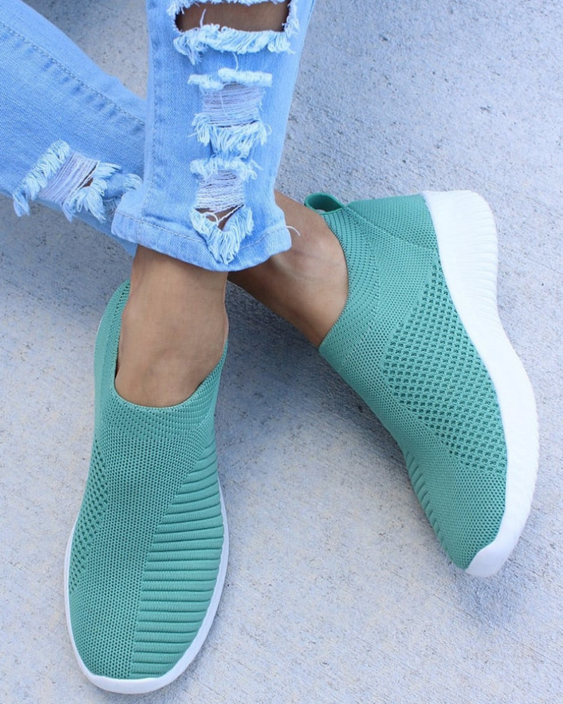 Green slip-on sneakers with white soles, featuring a breathable knit design, worn with ripped light blue jeans on a concrete surface.