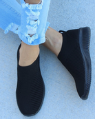 Black slip-on sneakers with textured knit design, worn with distressed light blue jeans. Comfortable, casual footwear for everyday style.