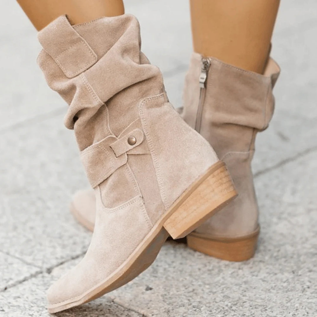 Beige suede ankle boots with slouchy design, wooden block heel, and side zipper on a textured gray pavement. Fashionable women's footwear.