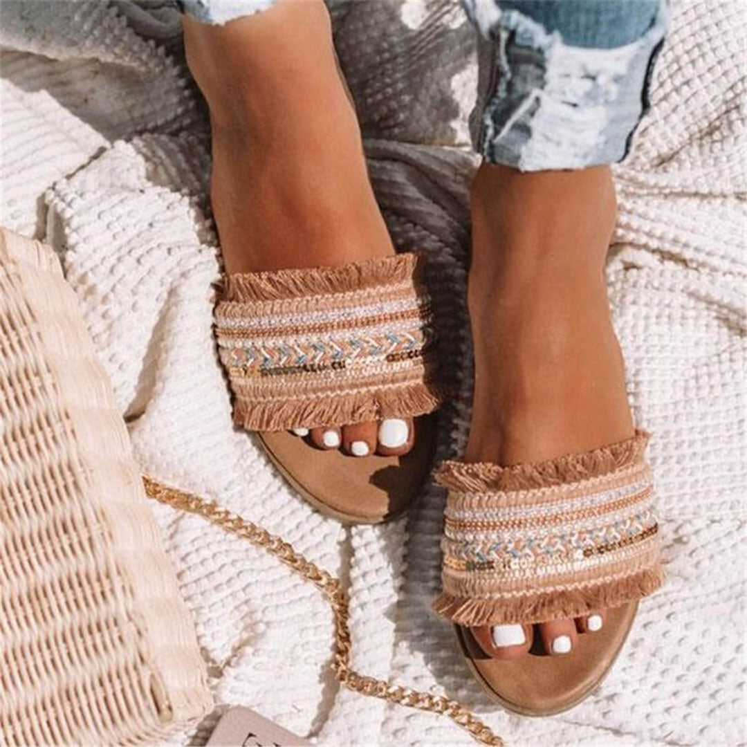 Beige woven sandals with fringe detail on a textured white blanket, featuring intricate embroidery, perfect for summer fashion and beachwear styling.