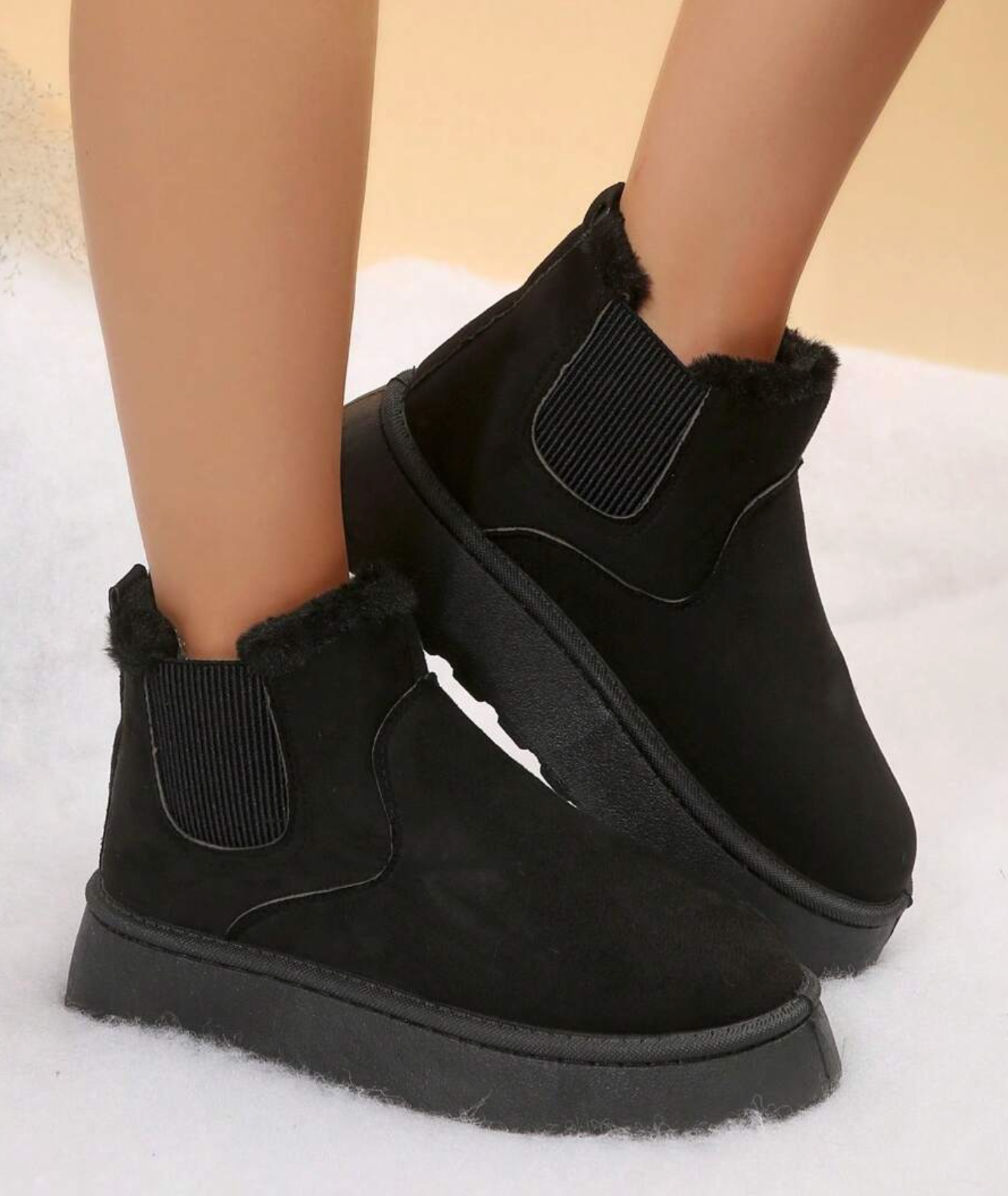 Black suede ankle boots with faux fur lining, elastic side panels, and platform soles on a snowy background. Winter footwear fashion.