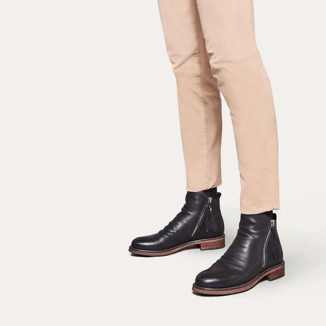 Black leather Chelsea boots with side zippers, worn with beige pants. Stylish men's footwear, perfect for casual and formal outfits.