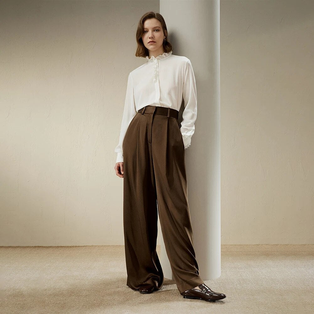 Woman wearing a white blouse and high-waisted brown wide-leg trousers, standing against a neutral background. Fashionable, elegant outfit for women.