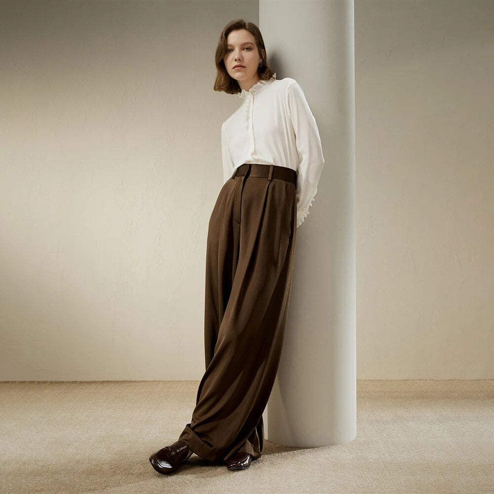 Woman in elegant white blouse and high-waisted brown wide-leg trousers, leaning against a pillar, showcasing modern minimalist fashion style.