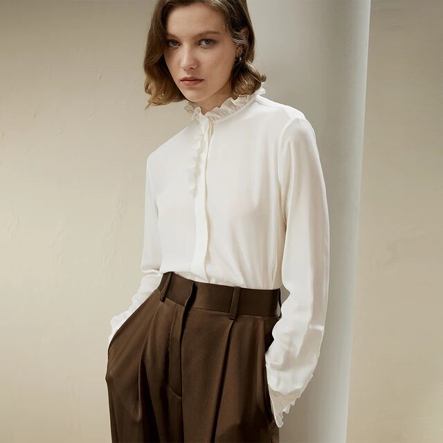 Woman wearing a white ruffled blouse and high-waisted brown trousers, standing against a neutral background. Fashionable, elegant women's clothing.