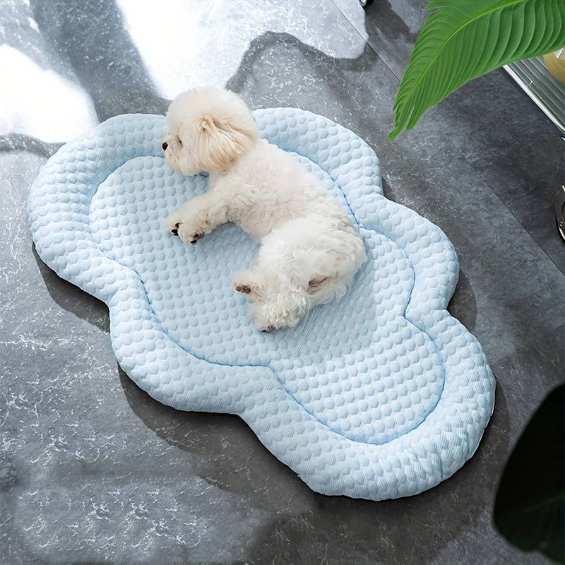 Small white dog resting on a cloud-shaped cooling mat for pets, placed on a gray marble floor, with a green plant leaf in the background.