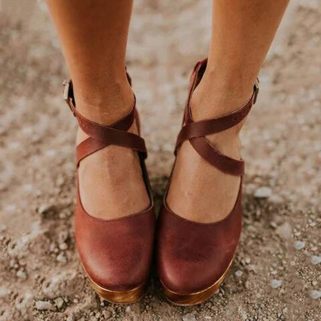 Brown leather cross-strap sandals on feet, standing on a textured dirt surface. Stylish women's footwear, comfortable, casual, summer fashion.