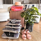 Rose gold suitcase with packing cubes, pink sandals, and a potted plant on a woven rug. Travel essentials for organized packing and stylish vacation.