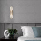 Modern black and gold wall-mounted reading light sconce on gray bedroom wall, above white bed with green pillow, next to decorative plant vase.