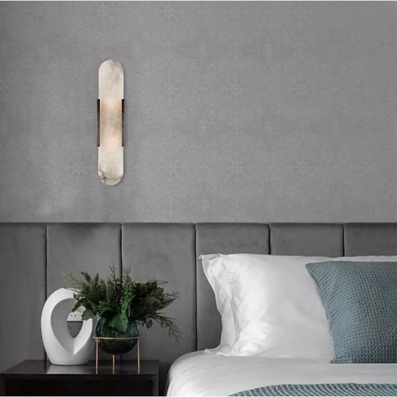 Modern black and gold wall-mounted reading light sconce on gray bedroom wall, above white bed with teal pillow and decorative plant on side table.