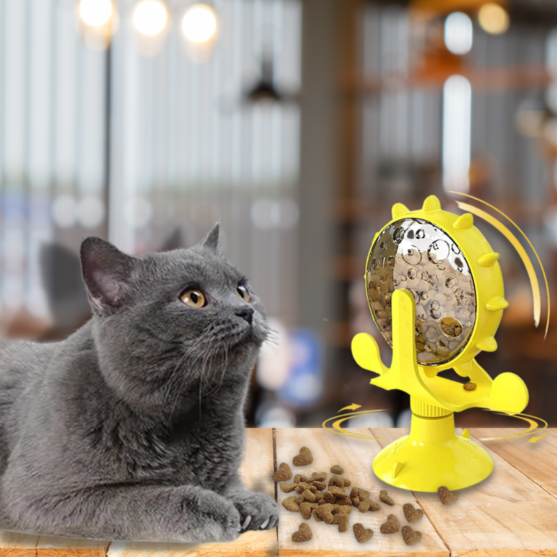 Gray cat with yellow interactive treat dispenser toy on wooden table, blurred background. Cat toy, pet enrichment, feline playtime.