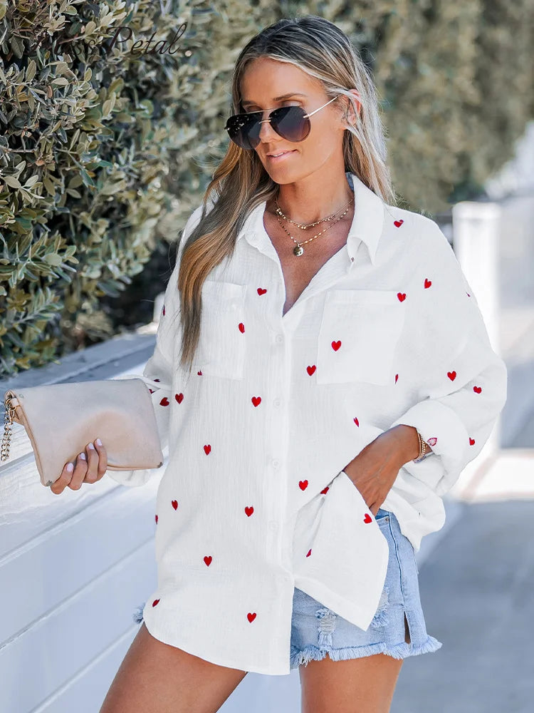 Woman wearing a white cotton embroidered heart crinkle woven shirt, casual loose long sleeve, paired with denim shorts, sunglasses, and holding a clutch.