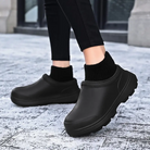 Black waterproof slip-on clogs with thick soles, worn with black socks on a snowy sidewalk. Durable winter footwear for outdoor activities.