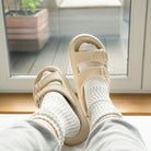 Beige sandals with double straps worn over white socks, resting on a windowsill. Cozy indoor footwear, casual style, comfortable home fashion.