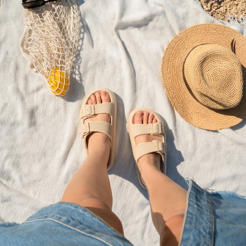 Person wearing beige sandals on a beach towel with a straw hat, denim shorts, and a net bag holding a lemon. Summer beach essentials, casual footwear.