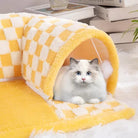 Fluffy white cat with blue eyes in a yellow and white checkered plush cat tunnel bed, surrounded by books and plants on a soft white rug.