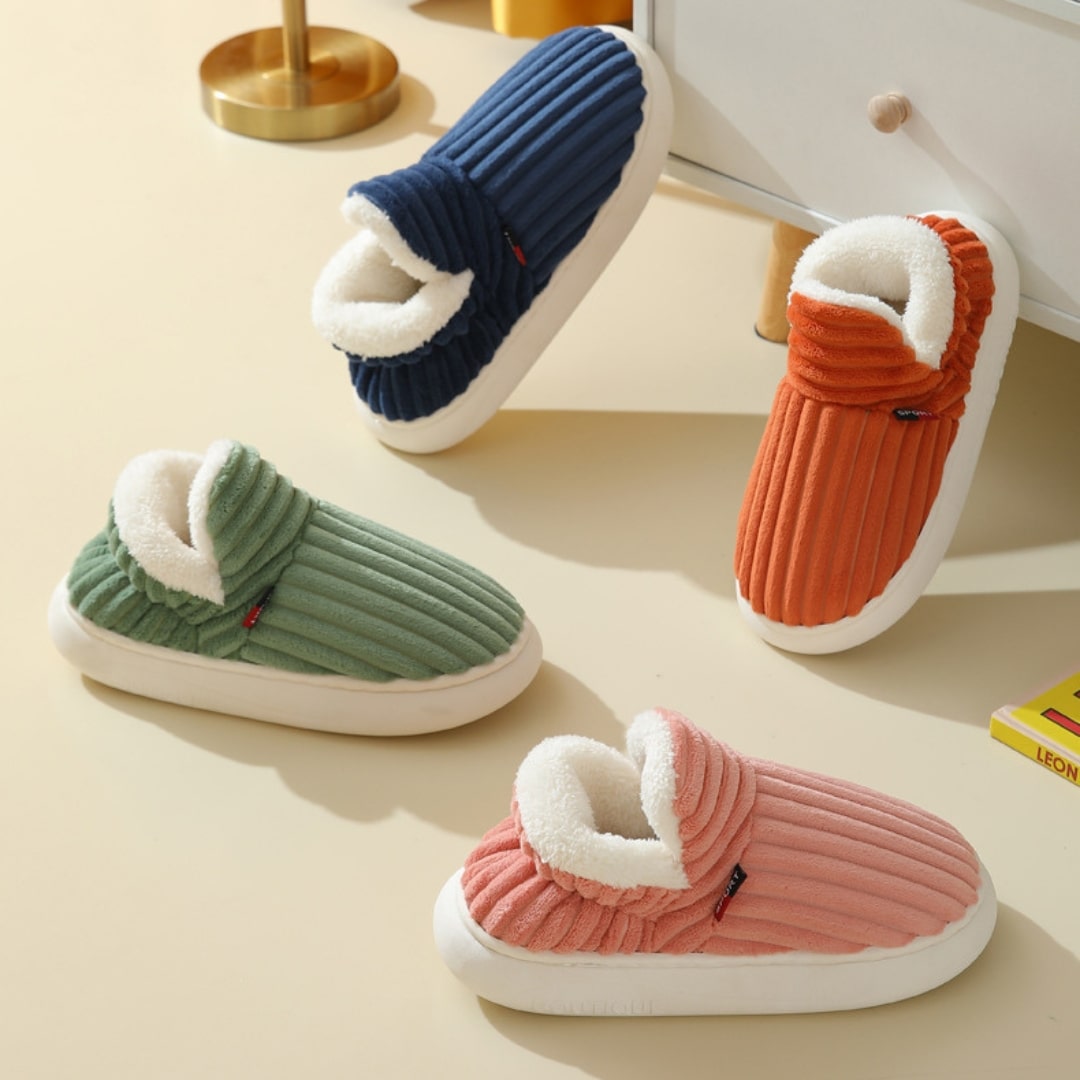 Cozy ribbed indoor slippers in blue, green, orange, and pink with plush lining, displayed on a beige floor near a white dresser and gold lamp.