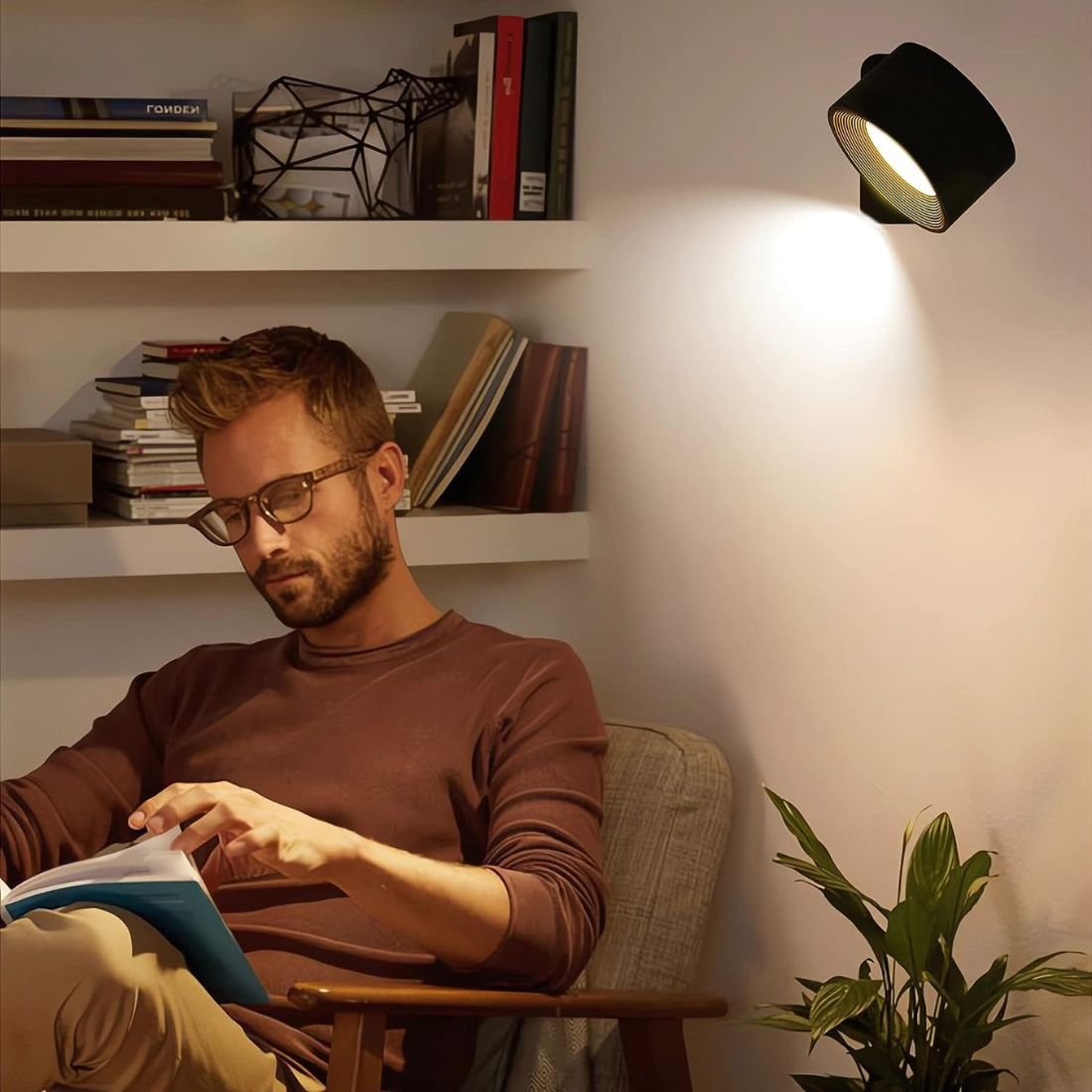 Man reading book under modern wall lamp in cozy living room; stylish lighting fixture, home decor, indoor plant, bookshelf background.