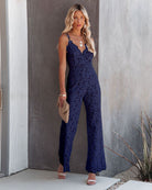 Woman in elegant navy lace jumpsuit, holding a beige clutch, standing against a modern concrete wall. Fashionable evening wear, stylish outfit.