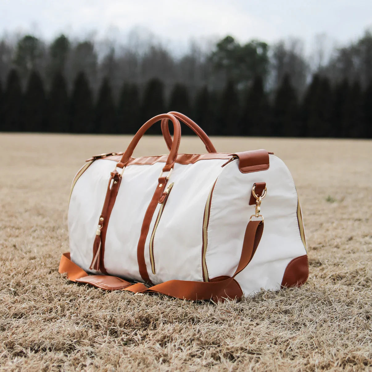 White and brown leather duffel bag on grass field, featuring gold zippers and handles. Perfect for travel, stylish luggage, foldable design.