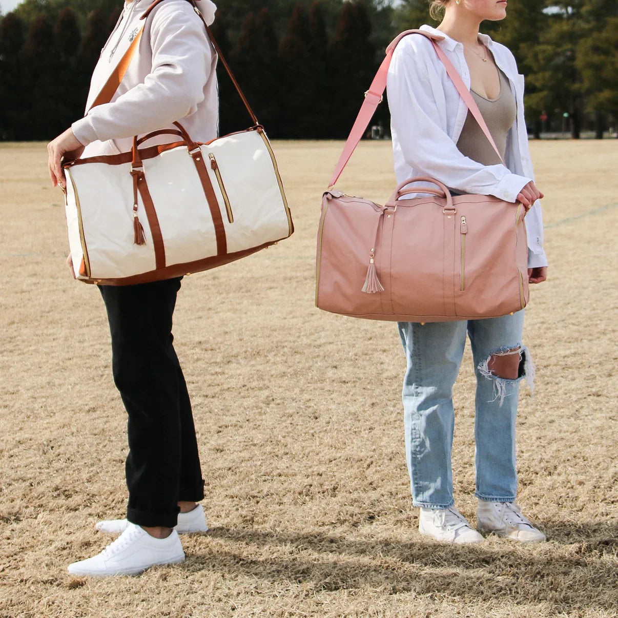 Two people standing outdoors holding stylish, large duffel bags; one white with brown accents, the other pink. Perfect for travel and gym use.