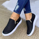 Black slip-on women's sneakers with breathable mesh design and white soles, paired with ripped jeans and a gold anklet. Casual spring-summer footwear.