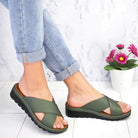 Women's green cross-strap sandals with cushioned sole, paired with rolled-up jeans, on a marble floor next to a white pot with pink flowers.