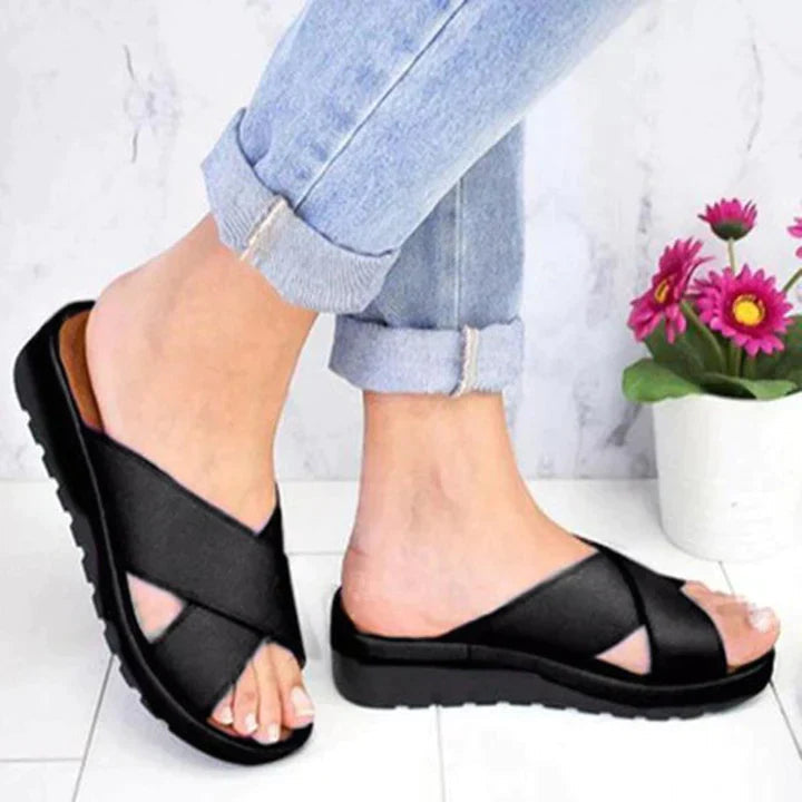 Black cross-strap sandals on feet, paired with rolled-up jeans, standing on a white floor. Comfortable footwear, casual style, women's fashion.