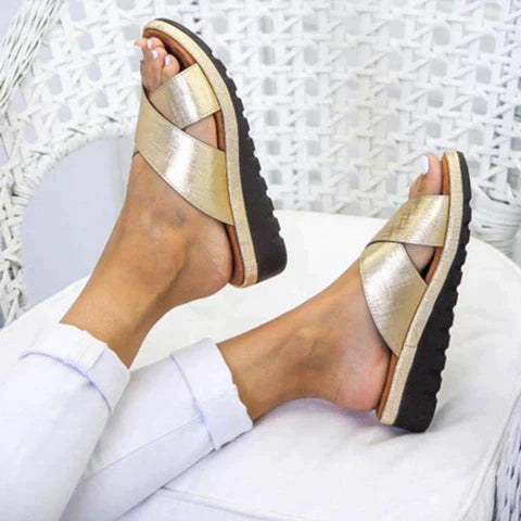 Gold metallic sandals with crisscross straps, black soles, and cork footbed, worn by a person in white pants, resting on a wicker chair.