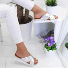 Silver platform sandals with crisscross straps on a woman's feet, paired with white pants. Modern footwear, stylish summer fashion, indoor setting.