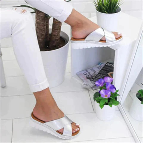 Silver slip-on sandals with crisscross straps and white platform soles, worn by a person in white pants, next to a potted plant and magazines.