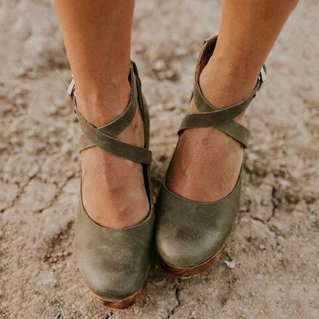 Women's green leather ankle strap shoes on cracked earth background, featuring cross-strap design and wooden soles. Fashionable footwear for casual wear.