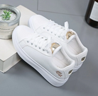 White leather sneakers with gold accents, featuring a rose embroidery on the side, placed on a gray surface next to a small potted plant.