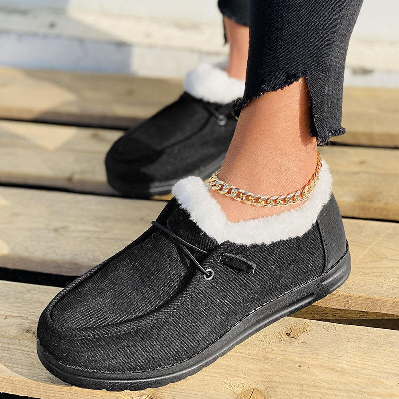 Black faux fur-lined moccasin slippers on wooden floor, featuring a gold anklet. Cozy winter footwear, stylish indoor shoes, comfortable slip-ons.