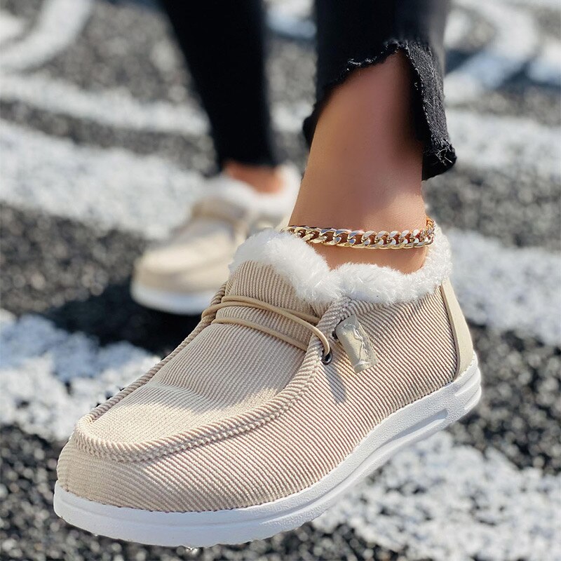 Beige corduroy slip-on shoes with white fur lining, worn with a gold chain anklet. Stylish women's casual footwear on a textured black and white background.