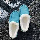 Turquoise corduroy slippers with plush white lining on textured black surface, cozy indoor footwear, warm winter slippers, stylish home comfort.