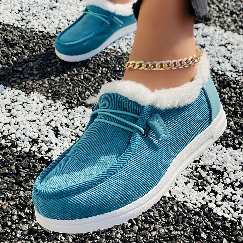 Turquoise corduroy loafers with white fur lining and white soles, worn on a textured black and white surface, featuring a gold ankle chain.