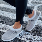 Women's casual gray slip-on shoes with white faux fur lining, worn with black jeans and a gold anklet, on a textured black and white surface.