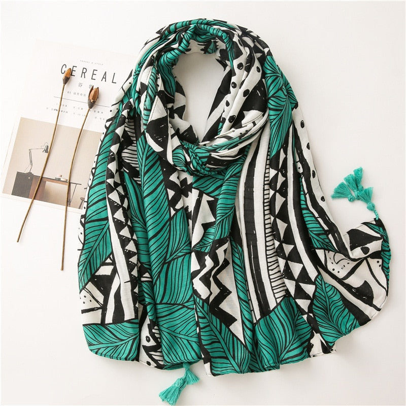 Green and black geometric patterned scarf with tassels, laid on a white surface next to a magazine and decorative sticks. Fashion accessory.