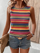 Woman wearing a colorful tribal print sleeveless top and denim shorts, accessorized with a woven handbag and jewelry, standing against a wooden backdrop.