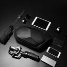 Black hardshell bag surrounded by tech gadgets, smartphone, tablet, smartwatch, sunglasses, umbrella, and gimbal on a dark surface.