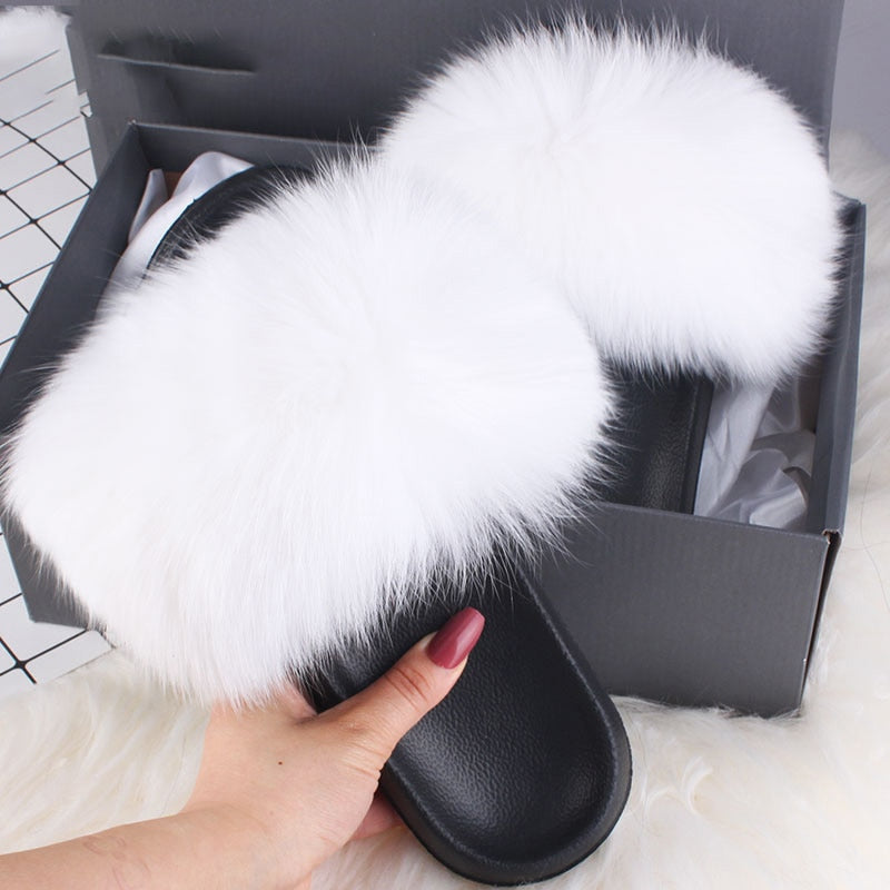 White fluffy fur slippers in a black box, held by a hand. Cozy, luxurious footwear, perfect for indoor comfort. Ideal for fashion-forward home lounging.