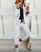 Woman in stylish white blazer and pants, black top, holding smartphone; casual chic fashion, modern women's outfit, trendy business casual look.