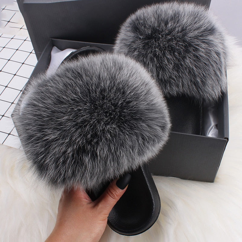 Gray faux fur slippers in a black box, featuring plush, fluffy design. Perfect for cozy indoor wear. Fashionable, comfortable footwear.