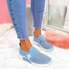 Women's blue slip-on sneakers with Velcro, enhancing posture, paired with frayed hem jeans, on a marble floor with a pink and white backdrop.