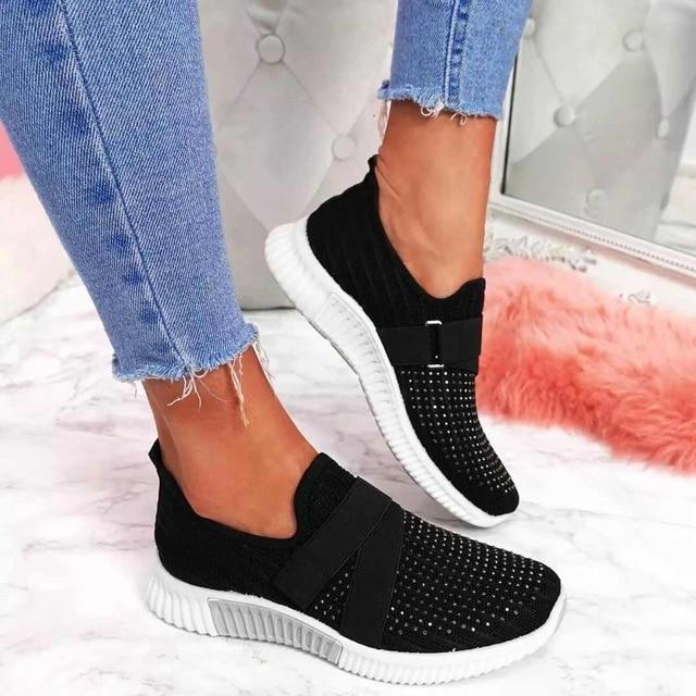 Black slip-on sneakers with Velcro strap, worn with blue jeans, on a marble floor. Comfortable women's footwear for improved posture.