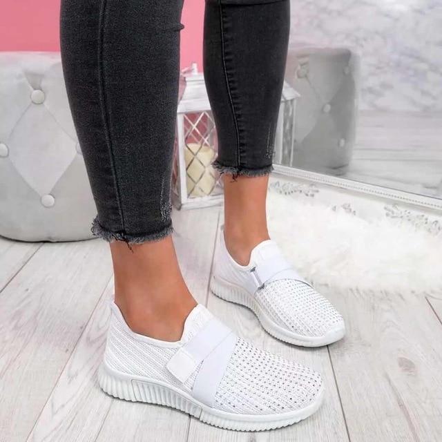 White slip-on sneakers with Velcro strap, worn by a person in black jeans, on a light wooden floor. Stylish women's footwear for improved posture.